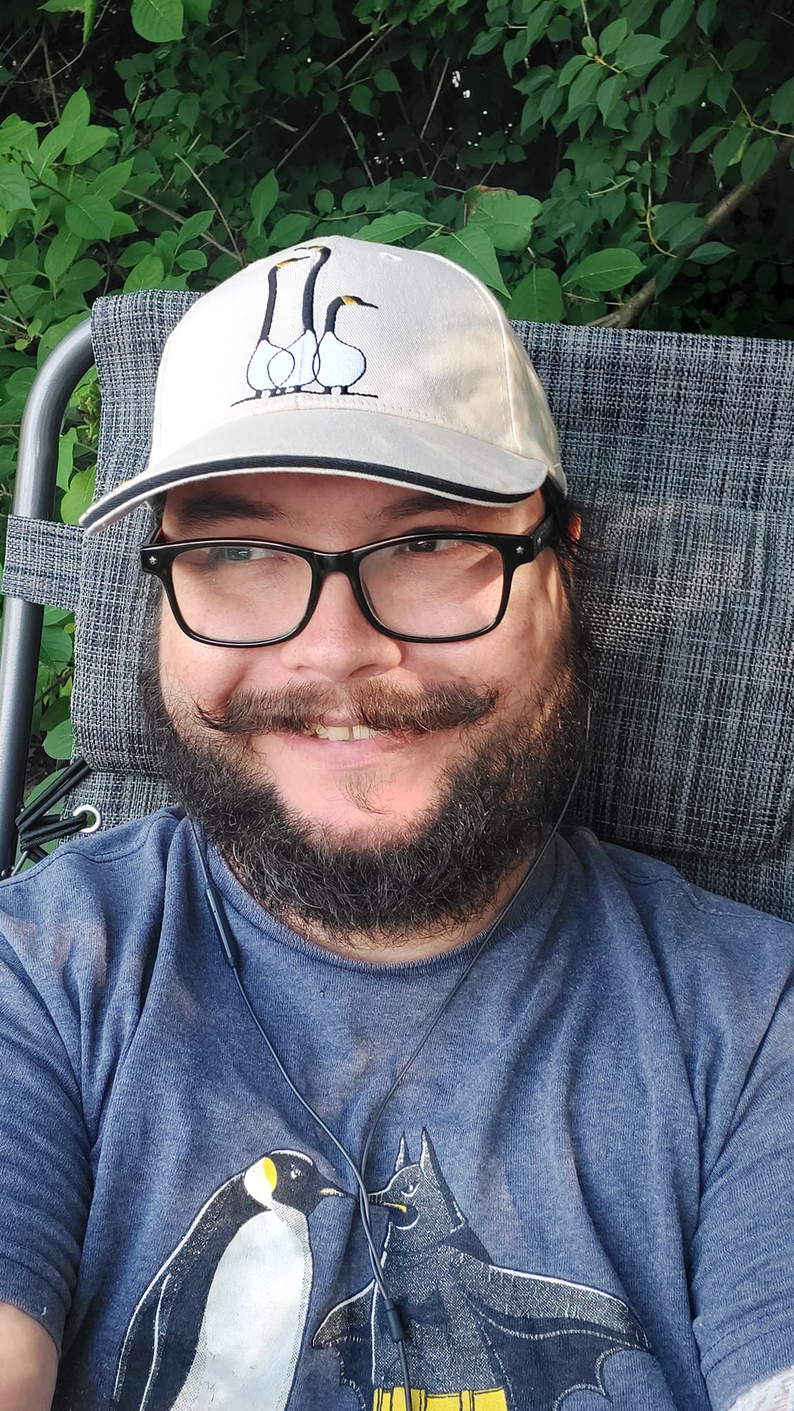 A man with short hair and bushy facial hair sits in a folding chair, smiing, with a hat with a goose on it